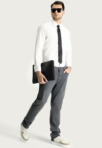 Hombre vistiendo una camisa blanca impecable, corbata negra, jeans grises y zapatillas grises y blancas, sosteniendo una funda de portátil negra con textura. Lleva unas gafas de sol negras.
