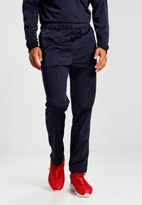Pantaloni athletic blu navy con vita elastica, gambe svasate e texture liscia, abbinati a scarpe da ginnastica rosse.
