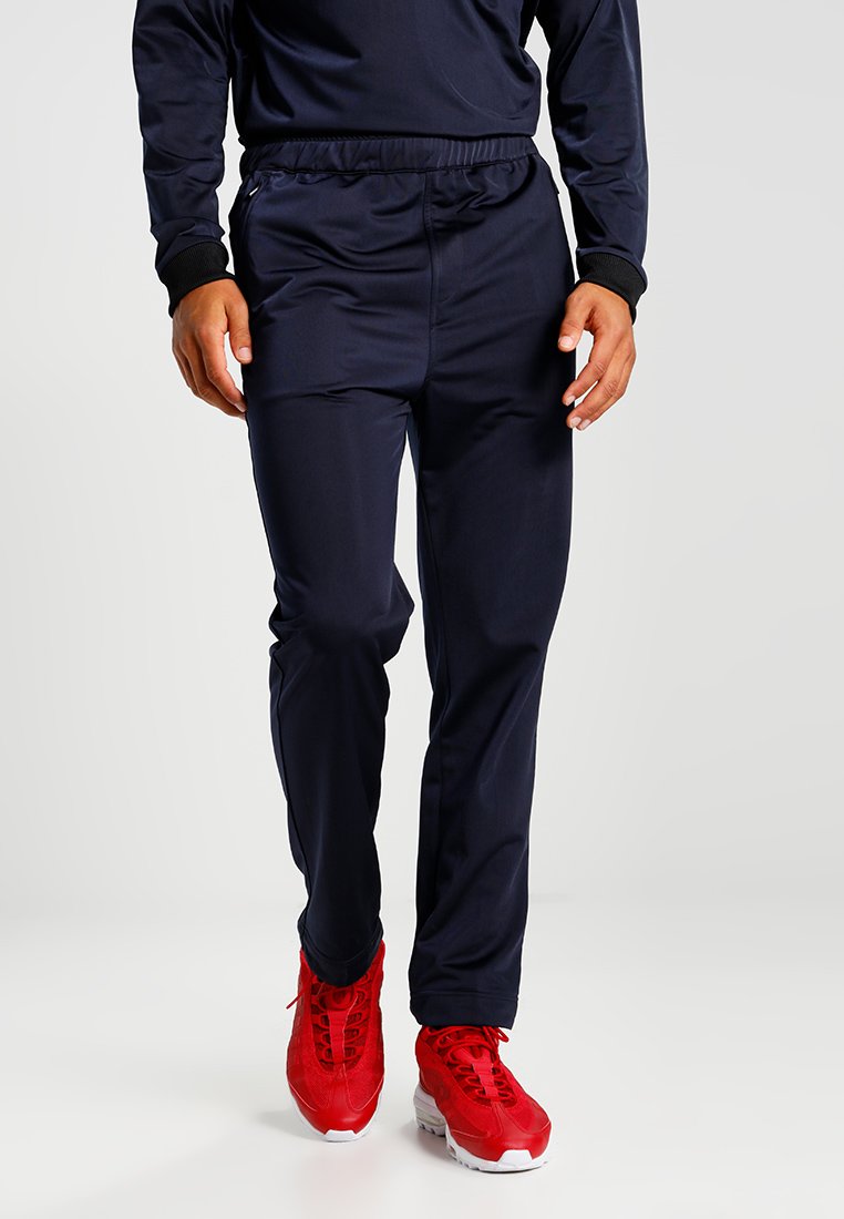 Pantaloni athletic blu navy con vita elastica, gambe svasate e texture liscia, abbinati a scarpe da ginnastica rosse.