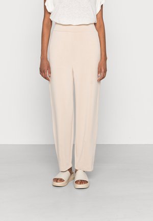 Osoba ubrana w beżowe spodnie o szerokich nogawkach, biały krótki top z krótkim rękawem oraz pasujące beżowe sandały na platformie z otwartymi palcami, stojąca na jednolitym tle.