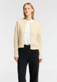 Cardigan lavorato a maglia in beige chiaro con motivo a rombi e bordi scallop. Presenta un colletto rotondo, chiusura con bottoni e maniche lunghe. Indossato sopra una camicia bianca.