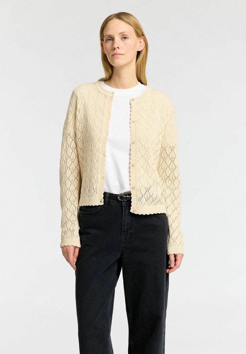 Cardigan lavorato a maglia in beige chiaro con motivo a rombi e bordi scallop. Presenta un colletto rotondo, chiusura con bottoni e maniche lunghe. Indossato sopra una camicia bianca.