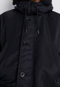 Chaqueta acolchada negra con cuello alto, botones de presión y cordones ajustables. Presenta una textura lisa y brillante y un forro interior acogedor.