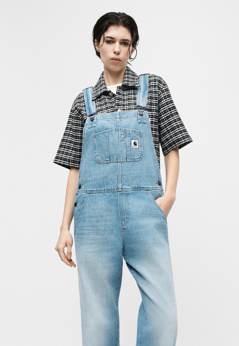 Jeune personne aux cheveux foncés portant une salopette en jean bleu clair et une chemise à manches courtes à carreaux noirs et blancs, debout avec une main dans la poche.