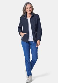 Marineblau gesteppte Jacke mit Stehkragen, Reißverschluss und zwei Seitentaschen, kombiniert mit blauen Jeans und hellen Turnschuhen.