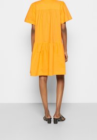 Robe en coton orange à manches courtes, dos froncé et jupe à volants. Texture lisse, longueur genou avec une coupe ample.