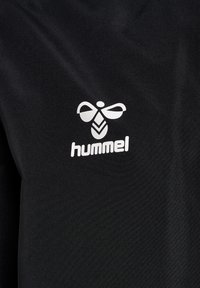 Tela negra con una textura brillante que presenta un logo blanco de "hummel" y un distintivo emblema de abeja, posicionado en el centro.