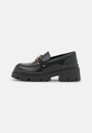 Zwarte leren loafer met stevige platformzool en gouden metalen gesp detail tegen een witte achtergrond.