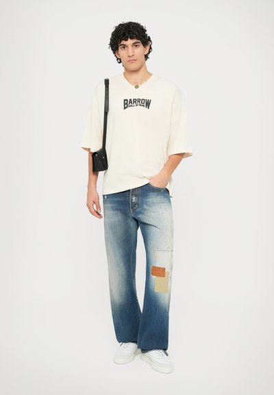 T-shirt graphique crème avec l'impression "BARROW HALL OF FAME", associé à un jean en denim usé avec des patches bleu clair et bleu foncé, des baskets blanches.