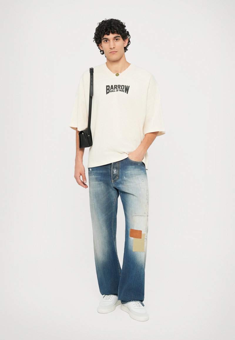 Cream graphic t-shirt with "BARROW HALL OF FAME" print, paired with distressed denim jeans featuring light and dark blue patches, white sneakers.