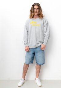 Grauer Sweatshirt mit gelbem "freestyle"-Text, kombiniert mit hellblauen Jeansshorts und weißen Turnschuhen, vor einem schlichten weißen Hintergrund.