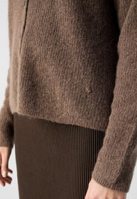 Cardigan en mélange de laine marron avec une finition texturée et cinq boutons, associé à une jupe côtelée marron. Insigne de marque notable à l'ourlet.
