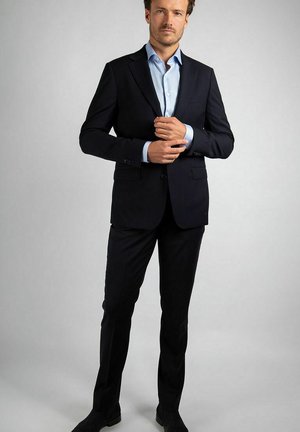 Man in een zwart pak en lichtblauw overhemd, die zijn manchetknopen verstelt, staande tegen een effen grijze achtergrond.