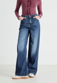 Wide-leg denim jeans in blue, worn high-waisted with a button closure and side pockets. The fabric has a slight fade with a smooth texture.
