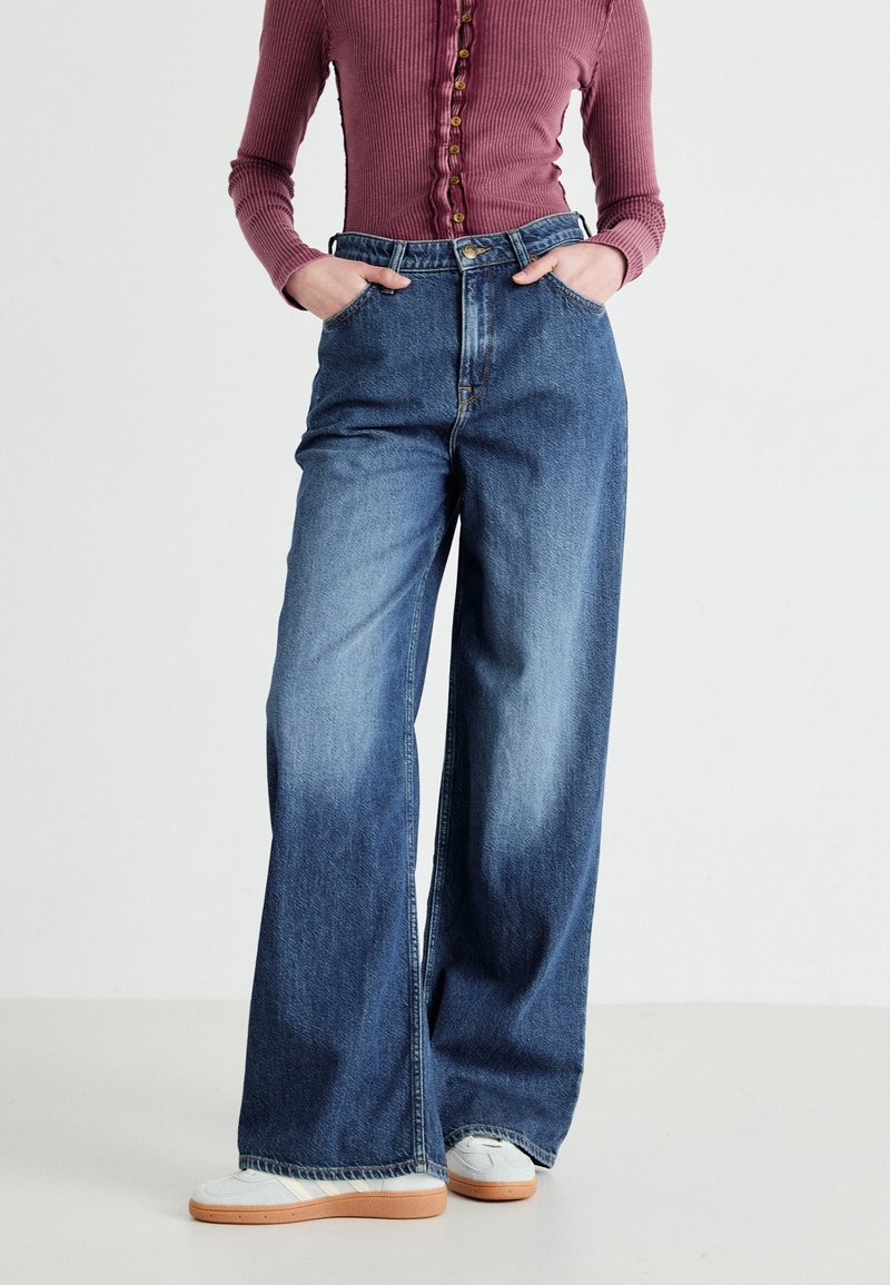 Wide-leg denim jeans in blue, worn high-waisted with a button closure and side pockets. The fabric has a slight fade with a smooth texture.