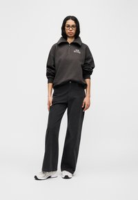 Σκούρο γκρι φούτερ με φερμουάρ και ψηλό γιακά, συνδυασμένο με μαύρο παντελόνι φαρδιάς γραμμής και λευκά αθλητικά παπούτσια.  Κείμενο λογότυπου στο στήθος του φούτερ.