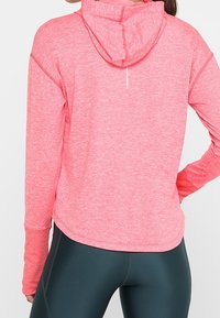 Hoodie sportivo rosa con un tessuto testurizzato. Presenta un cappuccio, maniche lunghe e un dettaglio di apertura sul retro. Abbinato a leggings scuri.