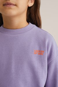 Lila Sweatshirt mit geripptem Rundhalsausschnitt, auf der linken Brust mit orangefarbener, gestickter Aufschrift "LUCK ON MY SIDE", weicher Baumwolltextur.