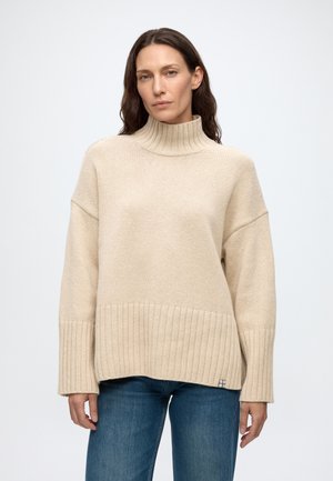 Femme aux longs cheveux bruns portant un pull à col roulé côtelé beige et un jean bleu, debout devant un fond uni clair.
