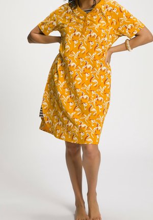 Frau trägt ein knielanges, senfgelbes Kleid mit kurzen Ärmeln und weiß-orangefarbenem Blumenmuster, posiert mit den Händen in die Taille gestützt.