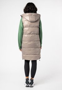 Osoba stoi tyłem, ubrana w długi beżowy puchowy kamizel, z kapturem, zielony sweter, czarne legginsy i czarne sneakersy za kostkę z białą podeszwą.