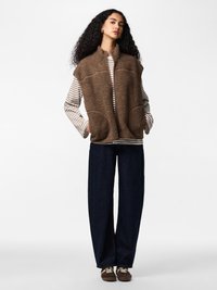 Göndör hajú nő barna sherpa mellényben, csíkos hosszú ujjú pólóval, sötét, bő szárú nadrágban és barna sportcipőben, fehér háttér előtt áll.