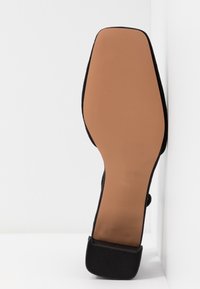 Chaussure à talon haut en tissu noir avec un talon texturé et une semelle lisse de couleur marron clair. La semelle est légèrement surélevée au niveau des orteils.