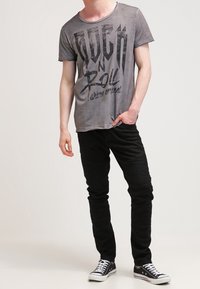 Camiseta gráfica gris con estampado de "Rock n Roll", combinada con pantalones negros de corte slim con acentos texturizados en las rodillas y zapatillas clásicas negras.