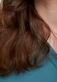 Capelli castano rossiccio con onde morbide, parzialmente visibili, accanto a un tessuto turchese con una texture liscia. Primo piano del contrasto tra capelli e tessuto.