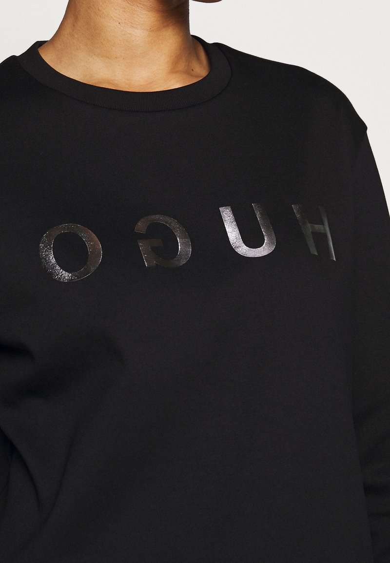 Schwarzer Pullover mit einem glänzenden, reflektierenden "HUGO"-Logo, das aufrecht über dem Brustbereich gedruckt ist. Weiches Baumwollmaterial.