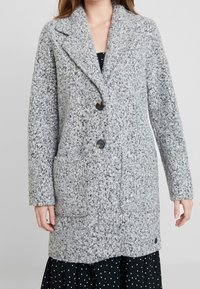 Harmaa teksturoitu takki, jossa on lovekaulus ja kaksi nappia. Siinä on päällystetyt taskut ja suora leikkaus, valmistettu paksusta, pörröisestä kankaasta.