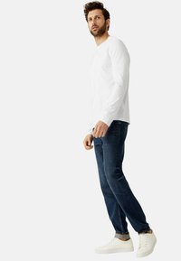 Witte lange mouwen shirt, donkerblauwe spijkerbroek en witte sneakers. De spijkerbroek heeft een klassieke pasvorm en het shirt heeft een ronde hals en een ontspannende textuur.