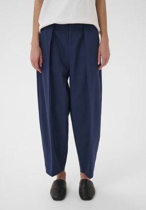 Person wearing loose-fitting navy pleated trousers, white top, and black slip-on shoes, standing against a plain background.