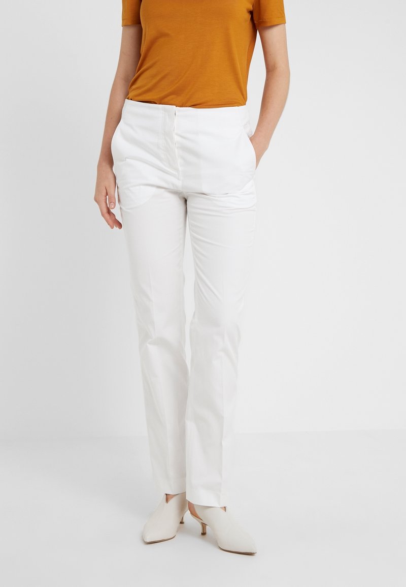 Bílé kalhoty s rovnými nohavicemi, se bočními kapsami a zapínáním na knoflík. V kombinaci s hnědým topem s krátkým rukávem a béžovými botami.