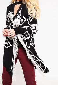 Cárdigan de patrón en blanco y negro con detalle de flecos, que presenta formas de diamante y un diseño largo y abierto en el frente. Combinado con pantalones burdeos.