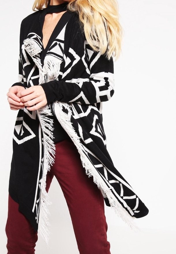 Zwart-wit gebreide cardigan met franjes, gekenmerkt door diamantvormen en een lang, open ontwerp. Gecombineerd met bordeauxrode broek.