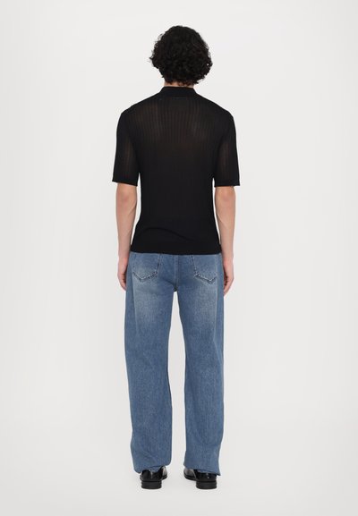 Fekete, rövid ujjú, bordás kötésű póló magas nyakkivágással, kék, bő szárú farmernadrággal párosítva. A hátulról készült nézet bemutatja a póló textúráját és illeszkedését.