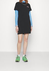 Robe t-shirt noire à manches courtes, avec un logo rose. Portée par-dessus un haut à manches longues bleu en côte. Associée à des baskets vertes et des chaussettes blanches.