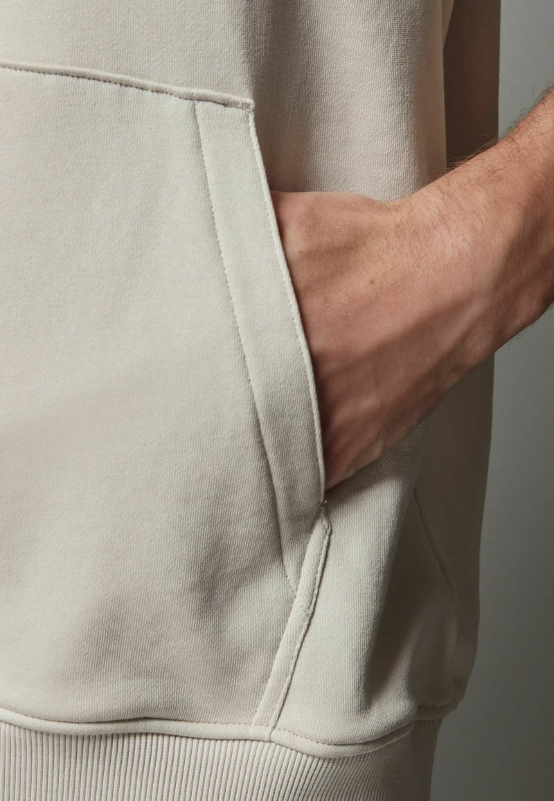 Beige hætte sweatshirt med en kengurulomme, ribbede kanter og blød tekstur. Pæne sømme og en afslappet pasform er synlige.
