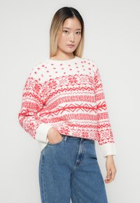 Mulher jovem com cabelo loiro liso, vestindo um suéter branco e vermelho com padrão e umas calças de ganga de cintura alta azuis, com uma mão no bolso.