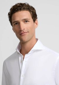 Camisa blanca de vestir con cuello extendido, con cierre de botones y una tela suave y texturizada. El modelo muestra una expresión neutra.