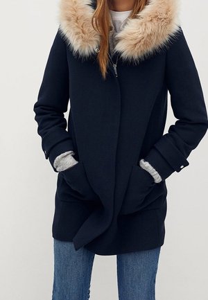 Kobieta w granatowym płaszczu z dużym beżowym kołnierzem z futra, szarymi dzianinowymi mankietami oraz niebieskich dżinsach, trzymająca ręce w kieszeniach płaszcza.