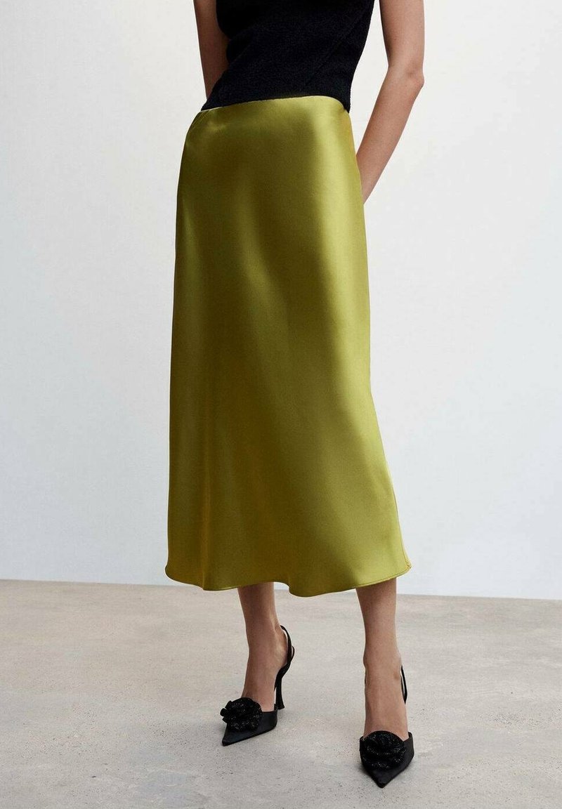 Femme portant une jupe midi vert olive brillante, un haut noir sans manches et des talons hauts noirs avec des nœuds décoratifs, debout sur un sol uni.