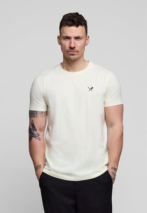 Mand med kort mørkt hår iført en lysebeige t-shirt med krydsede bestik-logo, sorte bukser og synlige tatoveringer på begge underarme.