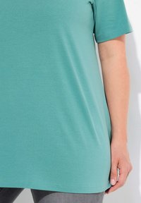 T-shirt corto a maniche corte di colore teal, realizzato in un tessuto morbido, con una vestibilità morbida e orlo curvo. Indossato con jeans grigi e una mano poggiata di lato.
