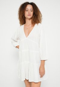 Weißes Stufenkleid mit tiefem V-Ausschnitt, langen Puffärmeln und strukturiertem Stoff. Verfügt über eine lockere Passform und einen gerafften Saum.