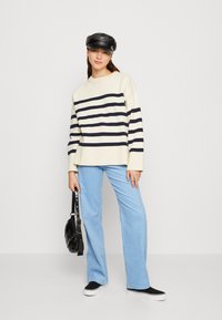 White sweater with navy horizontal stripes, paired with light blue corduroy pants and black sneakers. Accessories include a black cap and bag.