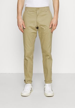 Mężczyzna noszący beżowe, dopasowane spodnie chinos z podwiniętymi nogawkami i białe sneakersy, stojący na jednolitym białym tle.