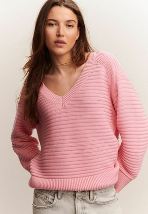 Rosa, ribbad stickad tröja med V-ringning och långa ärmar. Nederkantet har en bred, texturerad fåll. Bärs över ljusa jeans.