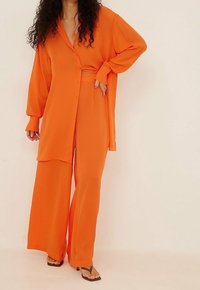Chemise orange à manches longues avec boutons, associée à un pantalon large en tissu lisse. Comprend des éléments matériels minimaux et une coupe décontractée.